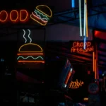 Papan neon berwarna cerah di pasar malam kota menampilkan iklan burger dan minuman, menggambarkan contoh kata kata iklan makanan yang menarik