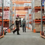 Dua pekerja mengelola inventaris barang di lorong gudang penyimpanan